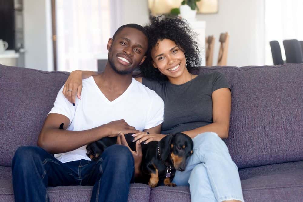 Happy couple sitting on couch at home cuddling their black and tan Dachshund 