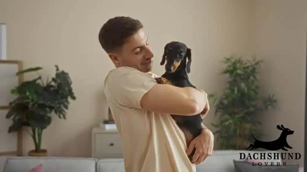 A smiling man in a beige shirt lovingly holds a small black dachshund in a cozy living room